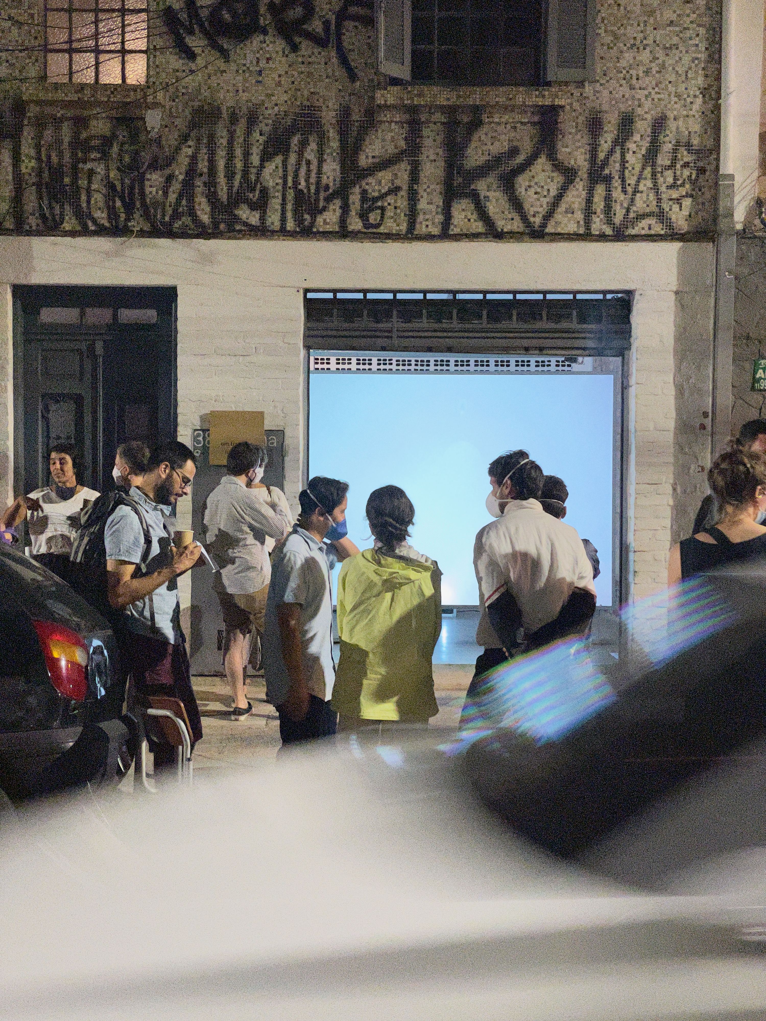 Marissa Lee Benedict and David Rueter, lâmina, 2021, Single-channel video installation, 2:00 TRT, Commissioned for the São Paulo project space um trabalho um texto (one work one text), lâmina is a one-night video work – a singular gesture – exhibited in parallel with a text by curator João Paulo Quintella. Documenting two hands, tentatively, awkwardly, bringing two pieces of 0.7 mm pencil lead (graphite) close enough to spark an electric carbon arc — an early form of spark lighting used for projecting films — before burning out, the work tightly loops together the history of moving image and its means of production.
This tight loop loosens over the course of the evening with a pile-up of strange and unexpected encounters: between the congenial atmosphere of the project space and the passing audience on the street; between the humming fan of a digital projector and the roar of traffic; between the loud squealing kisses of tiny graphite pencil lead rods, powered by a motorcycle battery, burning into light; and between the projected image of a beach at the mouth of a major colonial shipping channel, and the threshold of a garage door.
The exhibition contract for lâmina dictates that this version of the video (not including documentation of the video in situ) will only be publicly shown once (from sunset 18:04:00 to midnight on 26 September 2020 R. Rocha, 386 - Bela Vista, São Paulo, 013300-000, Brazil).
