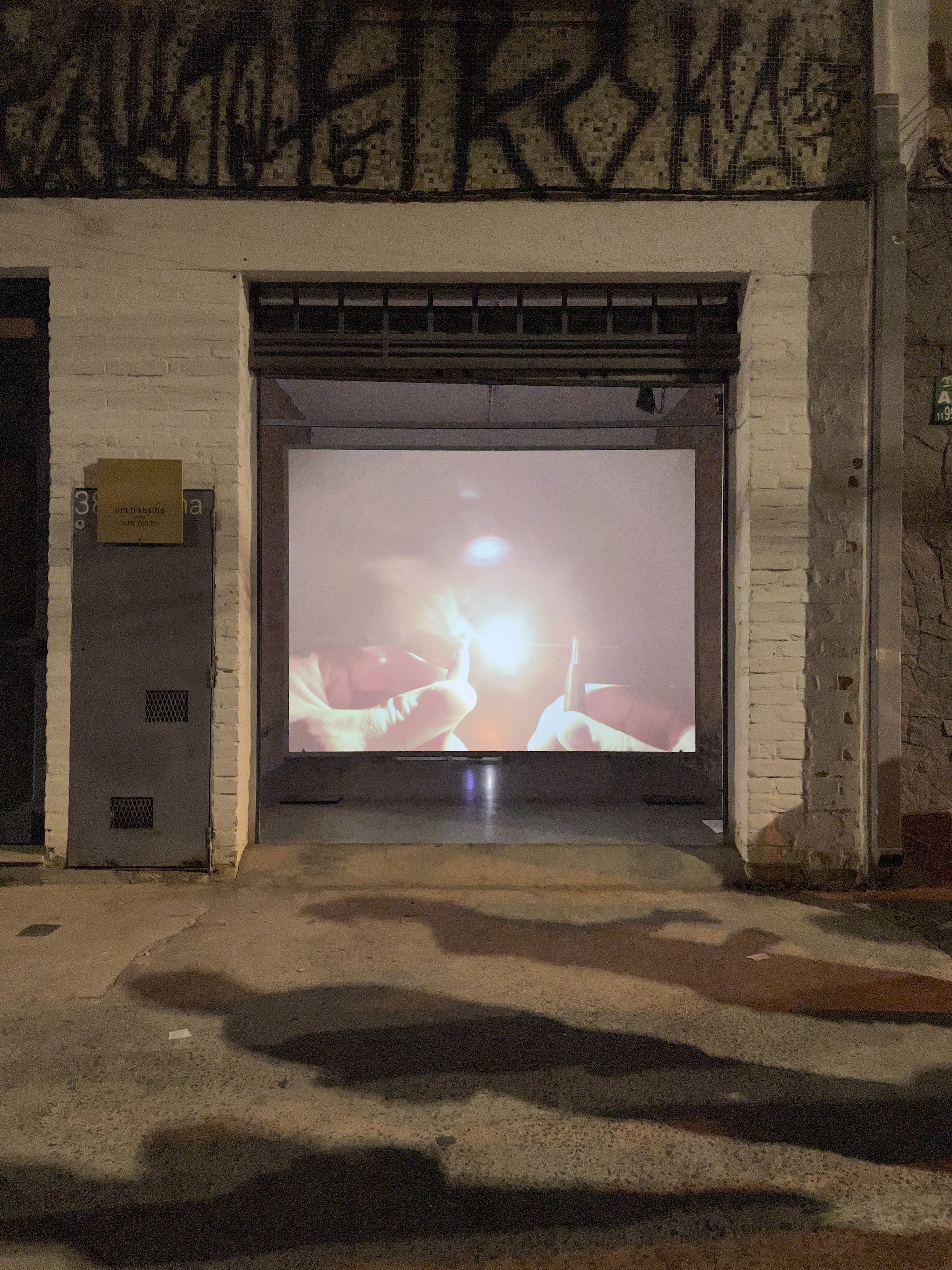 Marissa Lee Benedict and David Rueter, lâmina, 2021, Single-channel video installation, 2:00 TRT, Commissioned for the São Paulo project space um trabalho um texto (one work one text), lâmina is a one-night video work – a singular gesture – exhibited in parallel with a text by curator João Paulo Quintella. Documenting two hands, tentatively, awkwardly, bringing two pieces of 0.7 mm pencil lead (graphite) close enough to spark an electric carbon arc — an early form of spark lighting used for projecting films — before burning out, the work tightly loops together the history of moving image and its means of production.
This tight loop loosens over the course of the evening with a pile-up of strange and unexpected encounters: between the congenial atmosphere of the project space and the passing audience on the street; between the humming fan of a digital projector and the roar of traffic; between the loud squealing kisses of tiny graphite pencil lead rods, powered by a motorcycle battery, burning into light; and between the projected image of a beach at the mouth of a major colonial shipping channel, and the threshold of a garage door.
The exhibition contract for lâmina dictates that this version of the video (not including documentation of the video in situ) will only be publicly shown once (from sunset 18:04:00 to midnight on 26 September 2020 R. Rocha, 386 - Bela Vista, São Paulo, 013300-000, Brazil).
