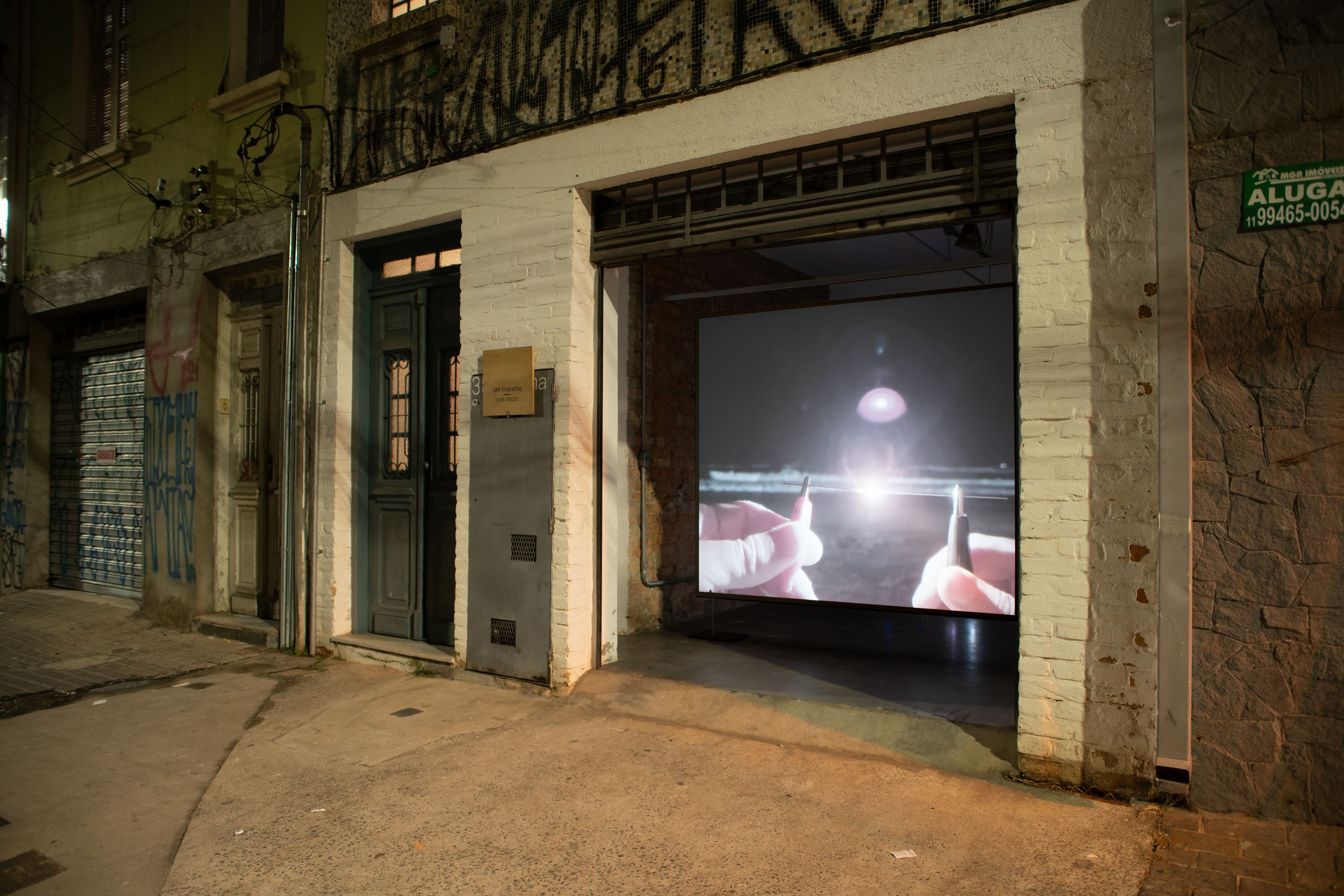 Marissa Lee Benedict and David Rueter, lâmina, 2021, Single-channel video installation, 2:00 TRT, Commissioned for the São Paulo project space um trabalho um texto (one work one text), lâmina is a one-night video work – a singular gesture – exhibited in parallel with a text by curator João Paulo Quintella. Documenting two hands, tentatively, awkwardly, bringing two pieces of 0.7 mm pencil lead (graphite) close enough to spark an electric carbon arc — an early form of spark lighting used for projecting films — before burning out, the work tightly loops together the history of moving image and its means of production.
This tight loop loosens over the course of the evening with a pile-up of strange and unexpected encounters: between the congenial atmosphere of the project space and the passing audience on the street; between the humming fan of a digital projector and the roar of traffic; between the loud squealing kisses of tiny graphite pencil lead rods, powered by a motorcycle battery, burning into light; and between the projected image of a beach at the mouth of a major colonial shipping channel, and the threshold of a garage door.
The exhibition contract for lâmina dictates that this version of the video (not including documentation of the video in situ) will only be publicly shown once (from sunset 18:04:00 to midnight on 26 September 2020 R. Rocha, 386 - Bela Vista, São Paulo, 013300-000, Brazil).
