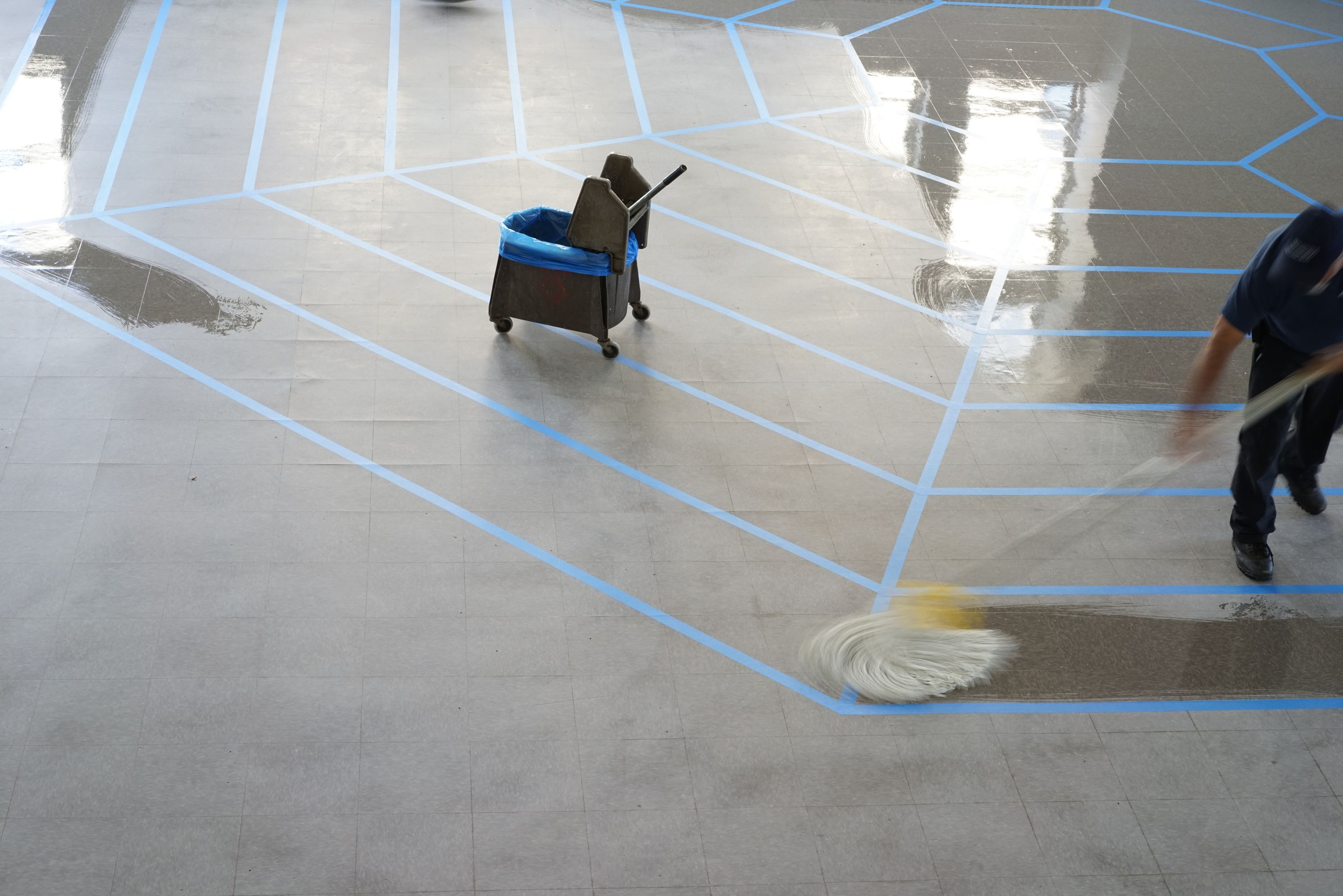 Marissa Lee Benedict, Daniel de Paula, and David Rueter, under cover of a solid object, 2022, Relief in floor wax, excerpt from the artists’ contract for the work deposition (2021), debris removed from ventilation ducts under the Chicago Board of Trade’s former corn futures trading floor, gelatin silver print (circa 1974, photographer unknown; Special Collections and University Archives, University of Illinois, Chicago)., In 2018, Rueter, Benedict, and de Paula acquired a used trading pit from the Chicago Board of Trade, now obsolete as trading goes digital. A huge octagon with seven tiered levels where traders bought and sold corn futures, the pit itself has since traveled to Sao Paolo and back to Chicago. Here it appears only as an outline at full scale, a ghost of itself that questions what can be distilled from a thing’s remnants. A pair of documents and a photograph appear like footnotes above the floor and in the opposite corner of the gallery.